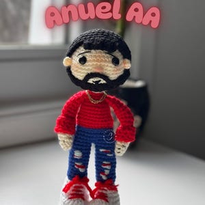 May include: A crocheted doll of a man with black hair and a beard, wearing a red shirt, blue jeans with rips, and white sneakers with red laces. The doll has a gold chain and the text "Anuel AA" above it.