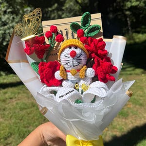 May include: A handmade crochet bouquet featuring a Doraemon character, red roses, and white lilies. The bouquet is wrapped in white paper and tied with a yellow ribbon. A golden butterfly and a card are also included.