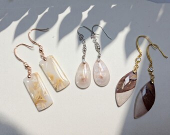 May include: A collection of four pairs of gemstone earrings. The earrings feature various shapes, including rectangular, teardrop, and leaf designs. The gemstones are in shades of white, beige, pink, and brown, with gold and silver-colored metal hooks.