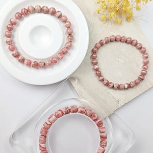 May include: Three pink and white striped gemstone bracelets. The bracelets are made of round beads and square beads. The bracelets are displayed on white and clear surfaces.