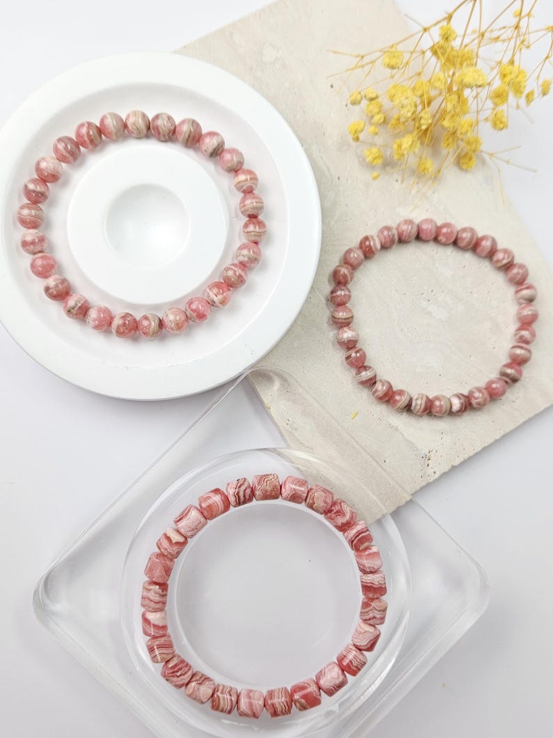 May include: Three pink and white striped gemstone bracelets. The bracelets are made of round beads and square beads. The bracelets are displayed on white and clear surfaces.