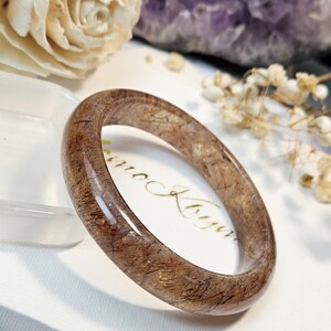 May include: A polished, circular bangle bracelet crafted from translucent brown rutilated quartz, featuring visible dark needle-like inclusions throughout the stone. The surface is smooth and reflective, showcasing the natural mineral patterns within.