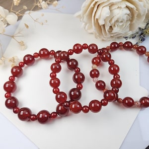 May include: Three red beaded bracelets with varying sizes of round beads. The bracelets are made of a translucent, reddish-brown material. The bracelets are displayed on a white surface with a white flower and a rose in the background.