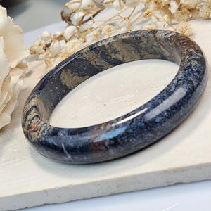May include: A polished stone bangle bracelet featuring a dark, mottled pattern of deep blue, charcoal gray, and earthy brown tones with visible mineral inclusions. The circular jewelry piece has a smooth, glossy finish and a rounded profile.