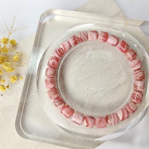 May include: A pink and white striped gemstone bracelet made of square beads. The bracelet is displayed on a clear square stand.
