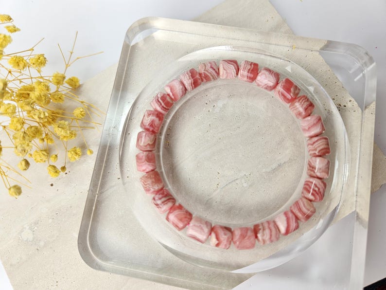 May include: A pink and white striped gemstone bracelet made of square beads. The bracelet is displayed on a clear square stand.