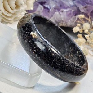 May include: A polished black stone bangle bracelet with a smooth, rounded surface and subtle, light-colored mineral inclusions throughout the dark material. The text Momokbymo is visible in the lower left corner of the image.