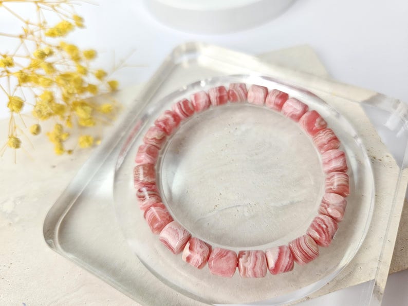 May include: A pink and white striped gemstone bracelet made of round beads. The bracelet is on a clear display stand.