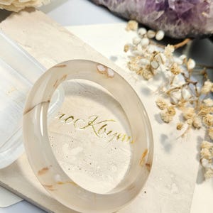 May include: A smooth, translucent beige bangle bracelet featuring subtle, natural brown and white inclusions throughout its polished surface. The circular accessory has a rounded profile and a glossy finish, showcasing the organic patterns within the stone.