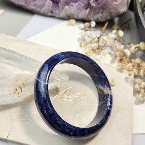May include: A polished, deep blue sodalite bangle bracelet featuring natural white and grey mineral inclusions throughout the stone. The circular band has a smooth, glossy finish and a rounded profile, showcasing the unique, mottled patterns of the stone.