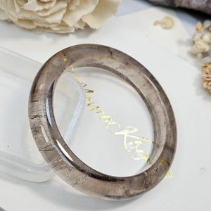 May include: A translucent, circular bangle bracelet made of smoky quartz with visible dark inclusions and subtle golden flecks throughout the stone. The polished, smooth surface of the bracelet reflects light, showcasing the natural greyish-brown color.