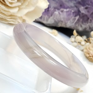 May include: A smooth, polished bangle bracelet crafted from translucent, light lavender-gray stone. The circular band features a rounded profile with a glossy, reflective surface that catches the light, showcasing the uniform, muted purple-toned coloration.