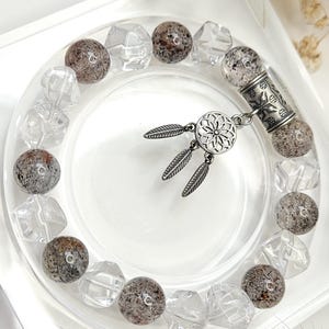 May include: A beaded bracelet featuring alternating clear, faceted beads and speckled, round beads. A silver-toned charm with a floral design and dangling feathers is attached. The bracelet is displayed on a clear, circular dish.