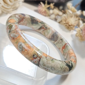 May include: A polished stone bangle bracelet featuring a marbled pattern with earthy tones of sage green, burnt orange, and cream. The smooth, rounded surface displays natural mineral inclusions and veins throughout the circular band.