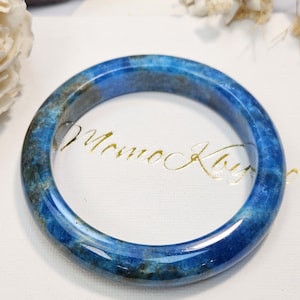 May include: A polished blue bangle bracelet with natural dark inclusions and a smooth, rounded surface. The bracelet features a vibrant, deep blue color with mottled patterns throughout the stone, displaying a glossy finish and a circular shape.