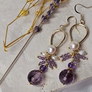 May include: Gold dangle earrings with a purple amethyst gemstone and white pearls. The earrings have a delicate, looped design.