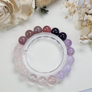 May include: A multi-colored beaded bracelet featuring a variety of gemstones. The bracelet includes pink, purple, and clear beads, with a clear plastic ring in the center. The bracelet is displayed on a white surface with floral accents.