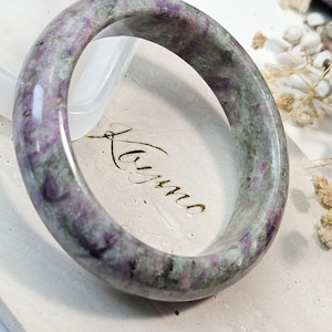 May include: A polished, circular bangle bracelet crafted from natural stone featuring a mottled pattern of deep magenta, grey, and white mineral inclusions. The smooth, rounded surface of the stone displays a glossy finish with visible natural variations.