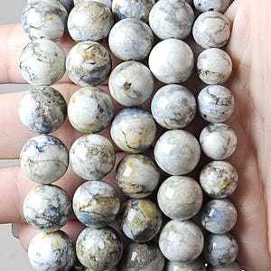 May include: A beaded bracelet featuring round, marbled beads in shades of white, gray, and tan. The beads are strung together, creating a bracelet. The bracelet is held in a hand, showcasing the natural stone texture and color variations.