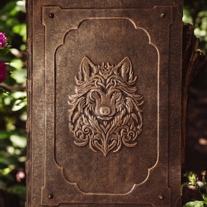 May include: A brown leather-bound journal with a detailed embossed wolf head design on the front cover. The wolf is surrounded by intricate floral patterns.