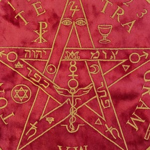 Puede incluir: Tela de terciopelo rojo con bordados dorados que representan un pentagrama con varios símbolos y texto. El diseño incluye una estrella central, un cáliz y una espada, con un borde circular que contiene símbolos y números adicionales.