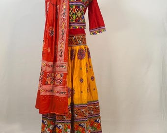 May include: A vibrant, traditional Indian outfit featuring a red and yellow color scheme. The ensemble includes a long-sleeved red top with intricate embroidery, a yellow skirt with detailed patterns, and a matching red scarf. The outfit is displayed on a mannequin.