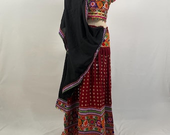 May include: A traditional Indian outfit, featuring a burgundy skirt with white dot accents and colorful embroidery. The top is a matching cropped style with intricate embroidery. A black shawl is draped over the outfit, adding a contrasting element.