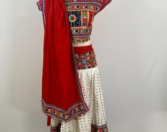 May include: A traditional Indian outfit, featuring a red top and matching skirt. The top is adorned with colorful embroidery and mirror work. The skirt is white with intricate patterns and embroidery. A red shawl is draped over the shoulder.