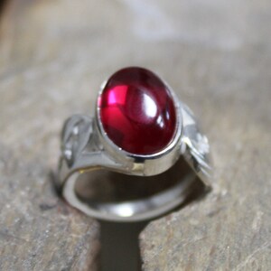 May include: A silver ring with a bright red oval gemstone set in the center. The ring has a unique, twisted design.