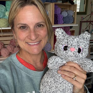 Pu&ograve; includere: Un giocattolo di peluche fatto a mano a forma di gatto, realizzato con filato soffice grigio e bianco maculato. L'animale di peluche presenta grandi occhi rotondi e scuri con bordi iridescenti e un piccolo naso rosa a tinta unita.