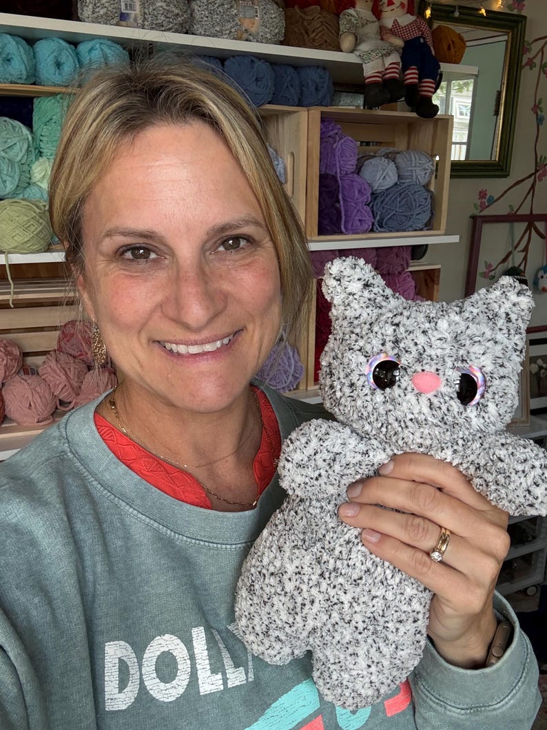 Pu&ograve; includere: Un giocattolo di peluche fatto a mano a forma di gatto, realizzato con filato soffice grigio e bianco maculato. L'animale di peluche presenta grandi occhi rotondi e scuri con bordi iridescenti e un piccolo naso rosa a tinta unita.