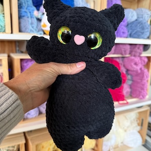 Pu&ograve; includere: Un gatto di peluche nero realizzato in filato morbido e testurizzato. Il gatto presenta grandi occhi rotondi e iridescenti nei toni del verde e dell'oro, un piccolo naso rosa chiaro a forma di cuore, orecchie a punta e un corpo arrotondato.