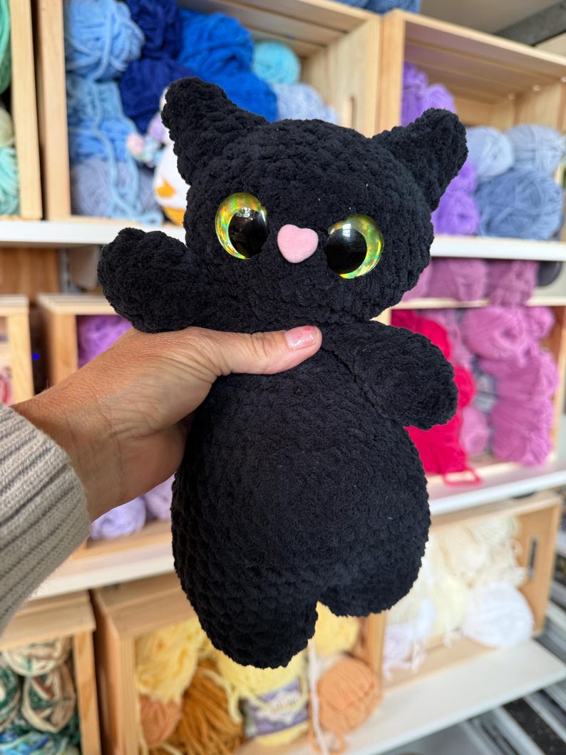 Pu&ograve; includere: Un gatto di peluche nero realizzato in filato morbido e testurizzato. Il gatto presenta grandi occhi rotondi e iridescenti nei toni del verde e dell'oro, un piccolo naso rosa chiaro a forma di cuore, orecchie a punta e un corpo arrotondato.