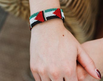 May include: A beaded bracelet with a pattern of red, white, green, and black beads. The bracelet is worn on a wrist. The bracelet's design is a repeating pattern of colors. The bracelet is a piece of jewelry.