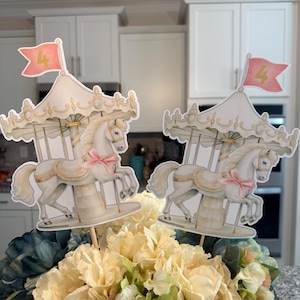 May include: Two white carousel horse centerpieces with pink flags that say "4" in gold. The horses are on white sticks and are in front of a bouquet of white and blue flowers.