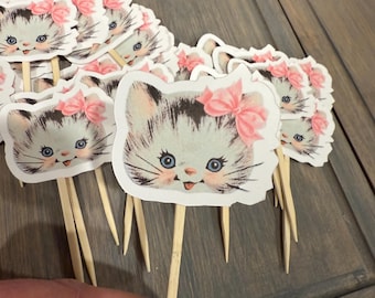 Op de afbeelding: Een set van 20 cupcake toppers met een schattig cartoon-kittens met een roze strik. De toppers zijn gedrukt op wit karton en bevestigd aan houten spiesjes.