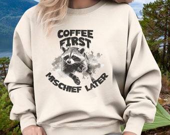 May include: A cream-colored sweatshirt with a black and white illustration of a raccoon. The text "Coffee First Mischief Later" is printed on the sweatshirt.