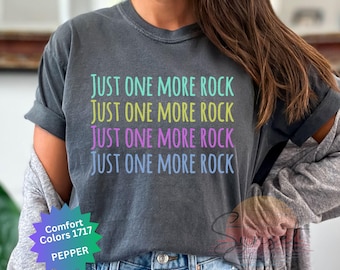 Puede incluir: Una camiseta gris oscuro con el texto "JUST ONE MORE ROCK" repetido cuatro veces en diferentes colores: turquesa, amarillo, rosa y azul.
