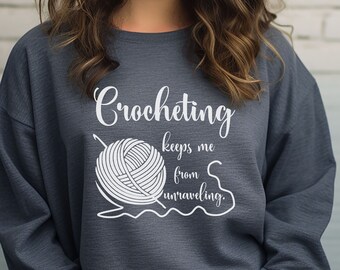 May include: A dark gray sweatshirt with the text "Crocheting keeps me from unraveling." in white. The text is accompanied by a white illustration of a ball of yarn and a crochet hook.