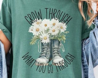 Puede incluir: Una camiseta verde con el texto "Grow Through What You Go Through" y un gráfico de un par de botas con margaritas que crecen.