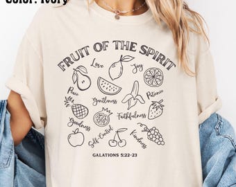 Może przedstawiać: Kremowa koszulka z napisem "FRUIT OF THE SPIRIT" i ilustracjami różnych owoców. Koszulka zawiera słowa takie jak "Miłość", "Radość", "Pokój" i "Życzliwość", a także odniesienie do Biblii Galacjan 5:22-23.