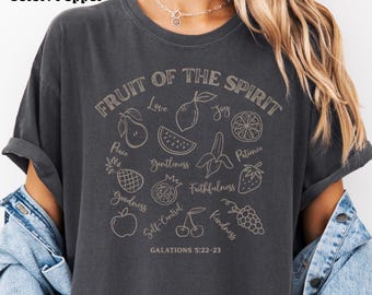 Może przedstawiać: Szara koszulka z napisem "FRUIT OF THE SPIRIT" i ilustracjami różnych owoców. Koszulka zawiera również tekst "GALATIONS 5:22-23" na dole. Projekt zawiera słowa takie jak "Miłość", "Radość", "Cierpliwość" i "Życzliwość".