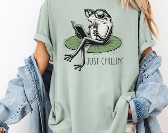 May include: A light green t-shirt featuring a cartoon frog wearing glasses and holding a book and coffee cup while sitting on a lily pad. The text "JUST CHILLIN'" is below the frog. The shirt is a solid color.
