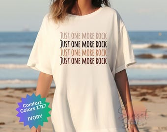 Puede incluir: Una camiseta blanca con el texto "JUST ONE MORE ROCK" repetido cuatro veces en diferentes tonos de marrón. La camiseta está hecha de una tela suave y cómoda y es perfecta para el uso diario.
