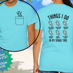 May include: A light blue t-shirt with a pocket on the front. The pocket has a giraffe peeking out of it. The back of the shirt has the text "THINGS I DO" and a series of giraffes with text describing activities related to giraffes. The text below the giraffes reads "IN MY SPARE TIME".