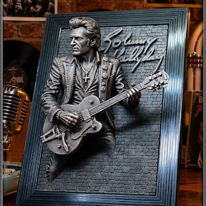 Pode incluir: Uma placa de parede tridimensional com uma escultura em relevo de cor prateada metálica de Johnny Hallyday tocando uma guitarra elétrica. A figura está sobre um fundo de tijolos escuros texturizados dentro de uma moldura retangular preta.