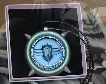 Puede incluir: Un collar con colgante plateado con un diseño azul y plateado. El colgante circular presenta un emblema central y cuelga de un cordón negro. El collar se presenta en una caja rosa.