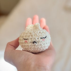 Peut inclure: Un jouet en peluche blanc en crochet en forme de petit pain avec un visage mignon et endormi. Le jouet a des yeux noirs, une petite bouche noire et des joues roses.