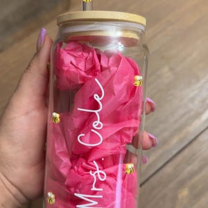 May include: A clear glass jar with a bamboo lid and a glass straw. The jar is filled with pink tissue paper and has the text "Mrs. Cole" written on it. There are small yellow bee decorations on the jar.