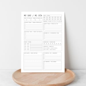 Op de afbeelding: Een zwart-wit dagplanner met Engelse en Spaanse secties voor taken, doelen en notities. De planner bevat secties voor stemming en hydratatie. Het ligt op een houten dienblad.
